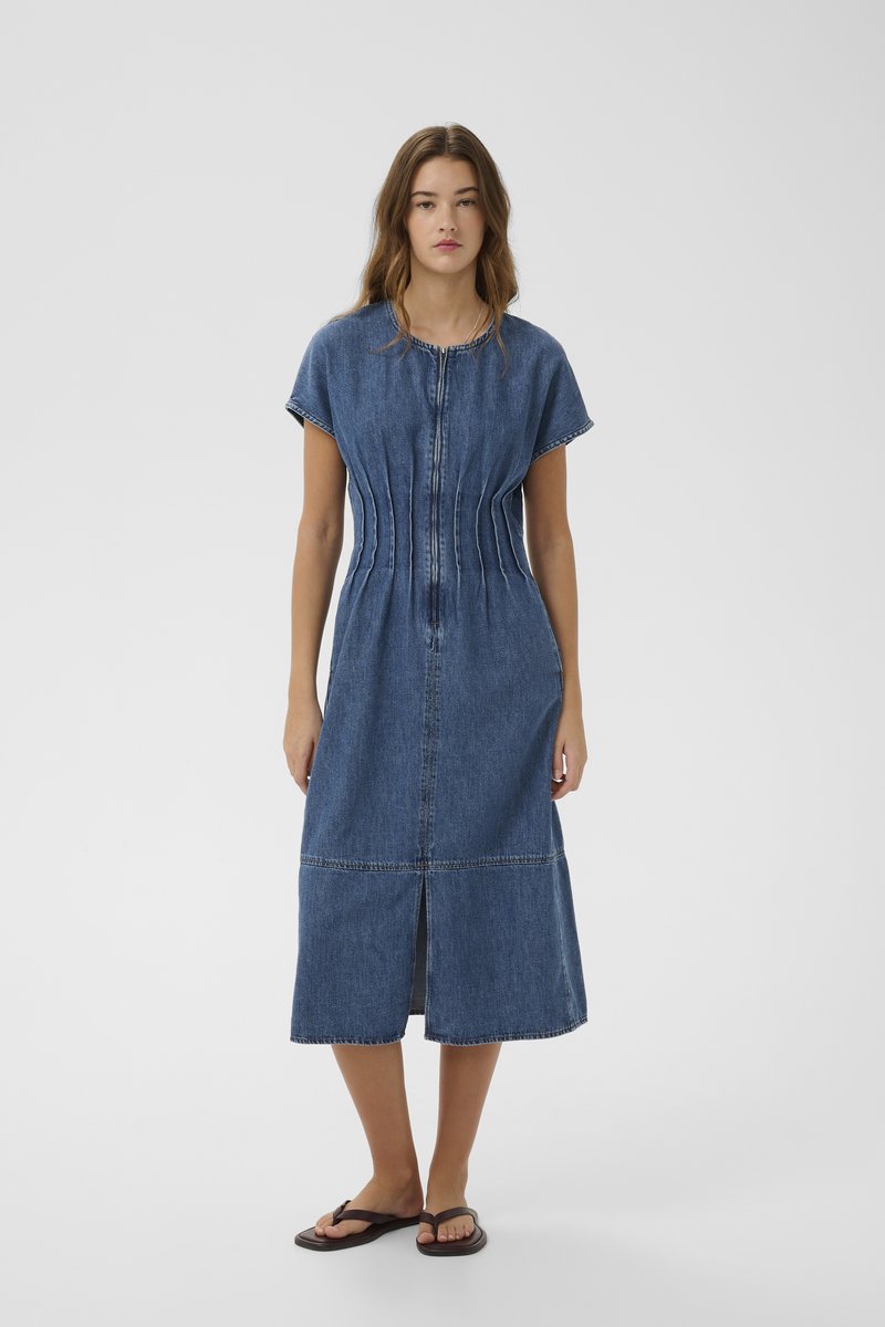 Femme portant une robe en jean mi-longue à manches courtes avec fermeture éclair devant, plis à la taille, fente frontale, et tongs foncées, debout devant un fond uni.