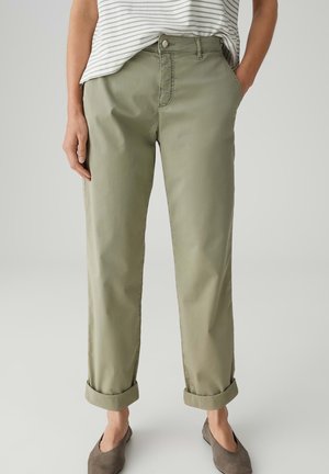 Frau trägt olivgrüne, hochgekrempelte Hose, gestreiftes, eingestecktes Hemd und braune Slipper, steht mit einer Hand in der Tasche.