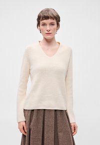 Κρέμα-χρώματος πλεκτό πουλόβερ με V-λαιμό, υφή υφάσματος και μακριά μανίκια, φορεμένο πάνω από μια καφέ φούστα με σχέδιο ψαροκόκαλο.