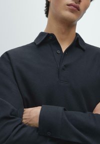 Schwarzes Poloshirt mit strukturierter Stoffqualität, drei Knöpfen am Kragen und langen Ärmeln. Die Arme des Models sind vor der Brust verschränkt.