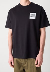 Osoba v černém tričku s krátkým rukávem, na kterém je bílé čtvercové logo s nápisem "Barbour International South Shields England" na hrudi.