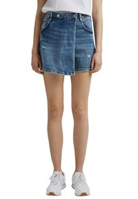 Jeans-Minirock in mittlerem Blau mit verwendetem Look, zwei Fronttaschen und vorderem Druckknopfverschluss. Kombiniert mit weißen Sneakers.