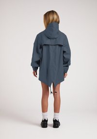Chaqueta impermeable azul con capucha, puños elásticos y una parte trasera más larga. Cuenta con cordones en el dobladillo para un ajuste ajustable. Textura suave.