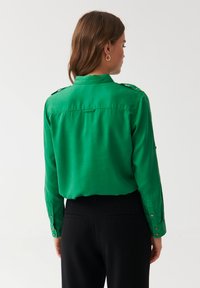 Cămașă verde din satin, cu o croială adaptată, având epoleți la umeri, mâneci lungi și manșete cu nasturi. Pantaloni negri vizibili la talie.