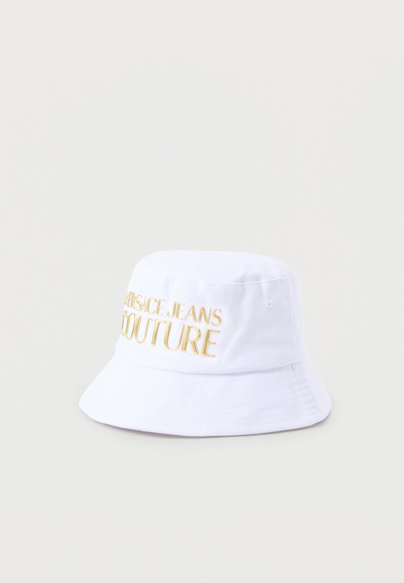 Λευκό καπέλο τύπου κουβά με χρυσή κεντημένη επιγραφή "Versace Jeans Couture" στο μπροστινό μέρος, απεικονισμένο σε απλό ανοιχτό φόντο.