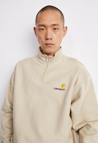 Bež dukserica s patentnim zatvaračem i dugim rukavima izrađena od mekanog materijala. Ima Carhartt logo u bordo i žutoj boji na lijevoj strani prsnog koša.