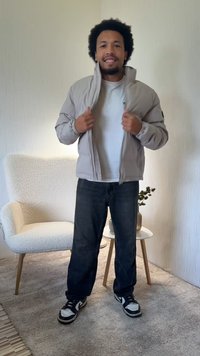 Lysegrå bomberjakke med lynlåslukning foran og elastiske manchetter, båret over en hvid t-shirt, kombineret med mørke jeans og sorte sneakers.