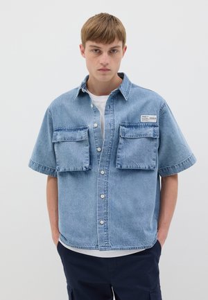 Camicia in denim azzurro chiaro a maniche corte con due grandi tasche sul petto, chiusura con bottoni e vestibilità comoda. Indossata sopra una t-shirt bianca.