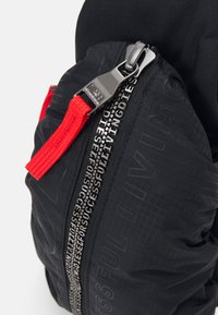 Sac zippé noir en tissu texturé avec un logo proéminent. Il dispose d'un zip rouge et de détails de couture visibles le long de la fermeture éclair.
