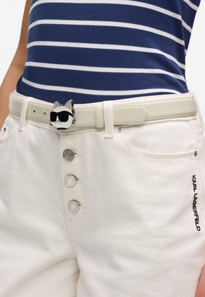 Beli pas z zaponko v obliki mačjega obraza, ki nosi črna sončna očala, oblečen v bele hlače z gumbno zadrgo in temno moder črtast top.