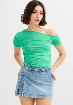 Joven con una blusa verde sin hombros y una minifalda de mezclilla azul, posando con las manos detrás de la espalda contra un fondo blanco.