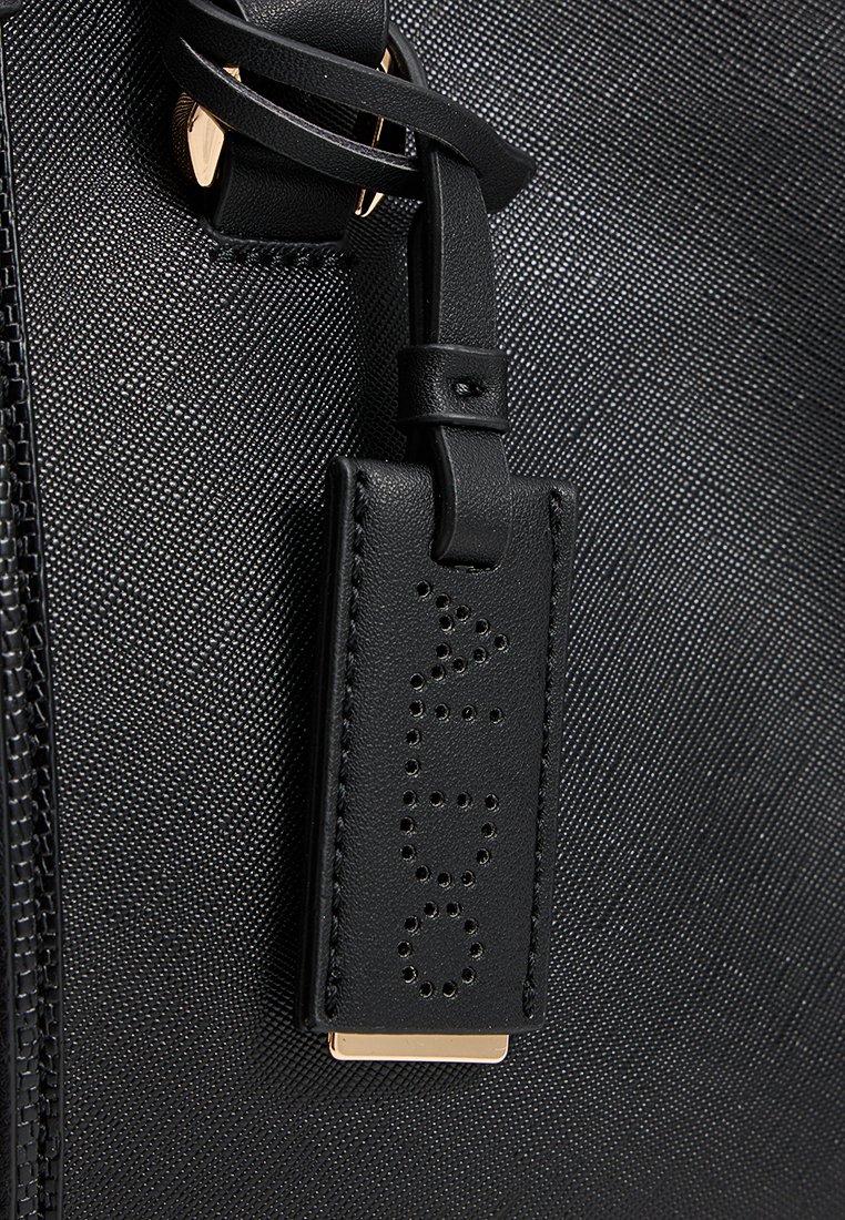 Borsa nera con texture caratterizzata da un'etichetta in pelle removibile con lettere perforate, fissata da una tracolla in pelle e dettagli in metallo dorato.