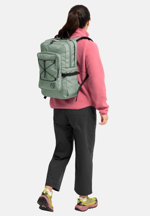 Person mit grünem Rucksack, rosa Fleecejacke, schwarzer Hose und gelben Wanderschuhen, stehend und von hinten auf weißem Hintergrund.