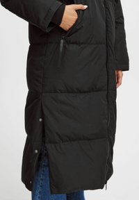 Čierna puffová bunda s dlhým dizajnom, obsahujúca bočnú vrecko, zips a patentky na leme. Hladký, izolačný materiál.