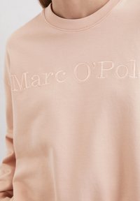 Helle rosa Sweatshirt mit Rundhalsausschnitt, ausgestattet mit erhabenen Text "Marc O'Polo" über der Brust. Glatte Textur, lässiges Design.