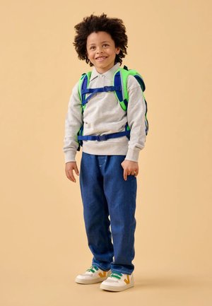 Gray sweatshirt, blue backpack with green accents, blue jeans, and white sneakers with green and yellow details. Smiling child stands on beige background.