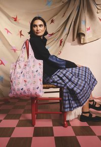 Ženska, oblečena v črni pulover in modro karo krilo, sedi na rdečem stolu s torbo sroza vzorca na zadnjem delu, z zvezdnatim ozadjem.