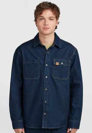 Junger Mann mit kurzen, lockigen Haaren, der ein dunkelblaues Jeanshemd mit zwei Brusttaschen vor einem einfarbigen Hintergrund trägt.
