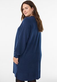 Femme aux longs cheveux bruns portant une robe tricotée à manches longues bleu marine, se retournant sur son épaule devant un fond blanc.