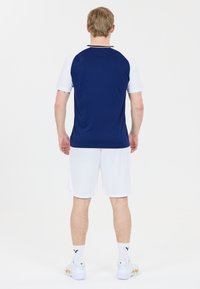 Herrarnas sportklädsel som består av en marinblå och vit kortärmad tröja, vita shorts och vita sneakers med gyllene detaljer.