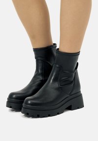 Botas de plataforma gruesa negras hasta la mitad de la pantorrilla, llevadas sobre piernas desnudas, que muestran una textura de cuero suave y una lengüeta en el talón.