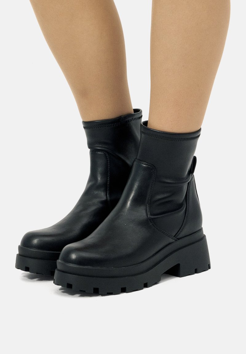 Botas de plataforma gruesa negras hasta la mitad de la pantorrilla, llevadas sobre piernas desnudas, que muestran una textura de cuero suave y una lengüeta en el talón.