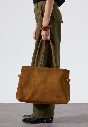 Henkilö, joka käyttää oliivinvärisiä housuja ja mustia kenkiä, pitää suuren ruskean mokkanahkaisen olkalaukun kahvoista sivullaan.