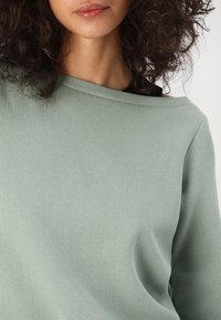 Maglione verde chiaro, scollato sulle spalle, con una texture morbida e colletto a coste. Presenta una vestibilità rilassata e maniche lunghe.