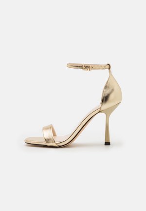 Złote metaliczne sandały na wysokim obcasie o minimalistycznym designie, z pojedyncim paskiem z przodu i regulowanym paskiem na kostkę. Gładkie wykończenie.