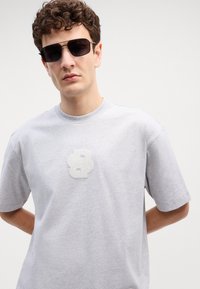 Sivá bavlnená tričko s krátkymi rukávmi, s fuzzy bielym logom "B" na hrudi. Model má tmavé slnečné okuliare a pózuje s rukami za chrbtom.