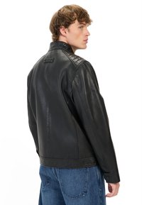 Zwarte leren jas met een hoge kraag, doorgestikte schouderaccenten en knoopmanchetten, gecombineerd met lichtblauwe denim broek.