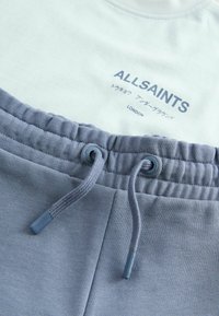 Grå bomullsjoggers med elastisk midja och dragsko. Ljusblå skjorta med "ALLSAINTS"-logotyp i marinblått tryck. Mjuk textur.