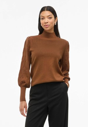 Pull en maille brun avec un col montant et des manches bouffantes, doté d'un motif texturé. Associé à un pantalon noir.