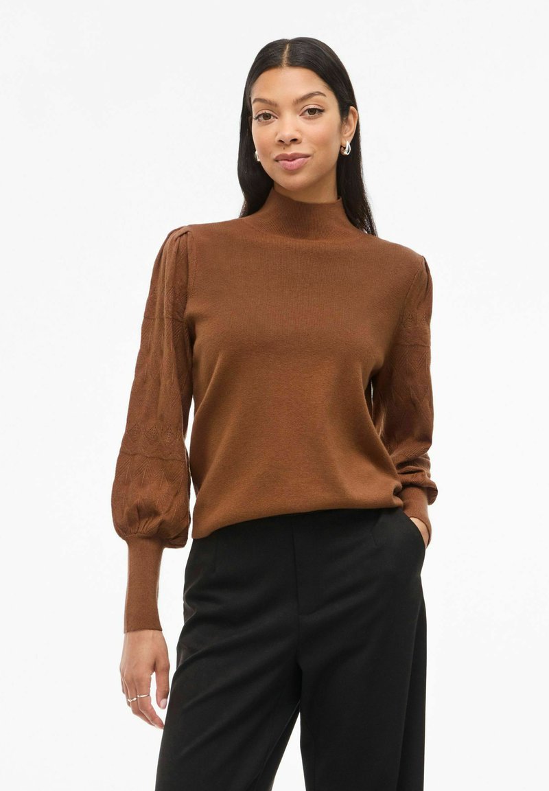 Pull en maille brun avec un col montant et des manches bouffantes, doté d'un motif texturé. Associé à un pantalon noir.