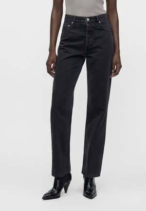 Model iført sorte højtaljede jeans kombineret med sorte støvler med spids tå og hæl samt en grå ærmeløs top, stående mod en ensfarvet baggrund.