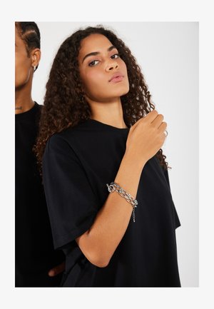 Camiseta negra de gran tamaño hecha de algodón, con mangas cortas, combinada con una pulsera de cadena de plata que presenta eslabones y un detalle de cierre.