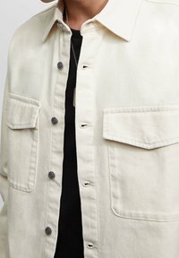 Chaqueta de mezclilla beige clara con cierre de botones en la parte delantera, dos bolsillos en el pecho y detalles en tono plateado. Tejido texturizado con cuello clásico.