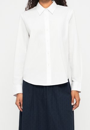 Personne portant une chemise blanche unie à manches longues boutonnée avec un col pointu et une jupe plissée foncée.