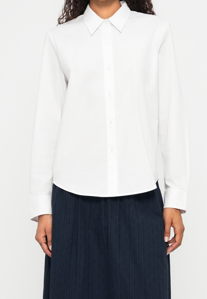 Personne portant une chemise blanche unie à manches longues boutonnée avec un col pointu et une jupe plissée foncée.