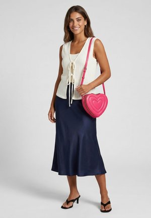 Smiling woman wearing white sleeveless tie-front top, navy midi skirt, black heeled sandals, and carrying a pink heart-shaped shoulder bag.