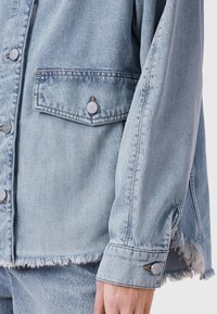 Šviesiai mėlyna džinsinė striukė su suskilinėjusiu apačiu, ilgomis rankovėmis, sagų sagtimis ir krūtinės kišenėmis. Lygus tekstūros paviršius su matomomis siūlių detalėmis.