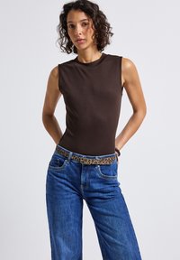 Frau mit lockigem Haar, die ein ärmelloses, dunkelbraunes Oberteil und eine Bluejeans mit einem Leopardendruckgürtel trägt, steht mit den Händen hinter dem Rücken.