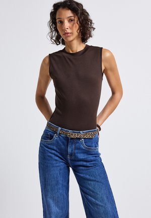 Femme aux cheveux bouclés portant un haut sans manches marron foncé, un jeans bleu avec une ceinture à motif léopard, se tenant avec les mains dans le dos.