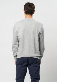 Hombre de espaldas vistiendo un suéter de manga larga gris claro y pantalones azul marino oscuros, frente a un fondo claro y sencillo.