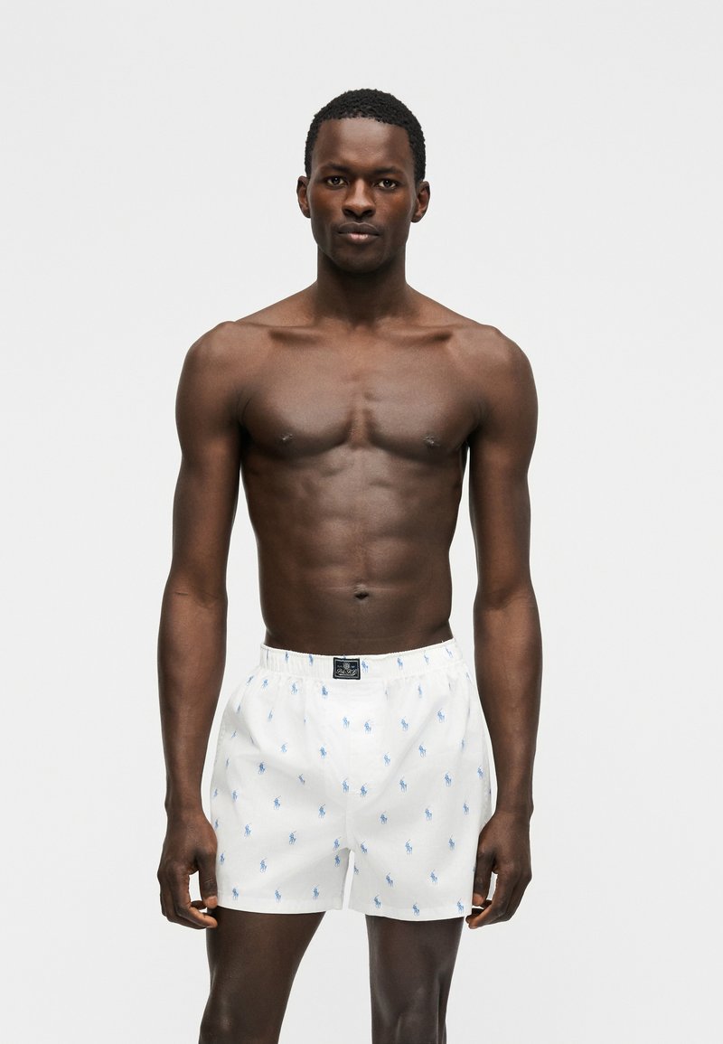 Gespiierde donkerhuidige man staand tegen een witte achtergrond, gekleed in witte boxershorts met blauwe geborduurde logo's.