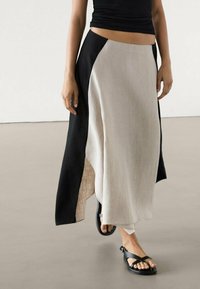 Zwarte en beige linnen rok met een hoge split en asymmetrisch ontwerp. Gecombineerd met zwarte sandalen, afgebeeld tegen een neutrale achtergrond.