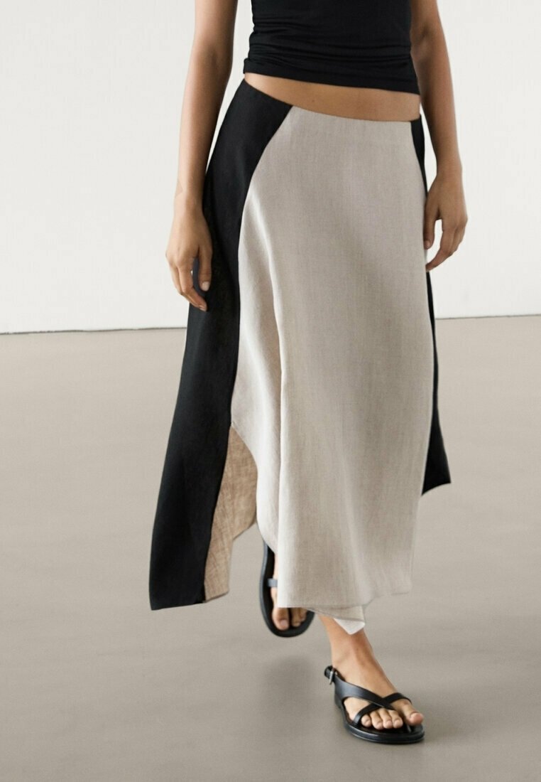 Zwarte en beige linnen rok met een hoge split en asymmetrisch ontwerp. Gecombineerd met zwarte sandalen, afgebeeld tegen een neutrale achtergrond.