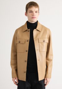 Κεραμιδί δερμάτινο μπουφάν με δομημένο σχέδιο, διαθέτοντας δύο τσέπες στο στήθος, κουμπώματα και γιακά με άκρες. Μαύρο τούλι κάτω από αυτό.