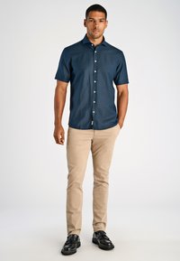 Man in een marineblauwe korte mouwen overhemd, beige broek en zwarte kwastloafers tegen een eenvoudige licht achtergrond.