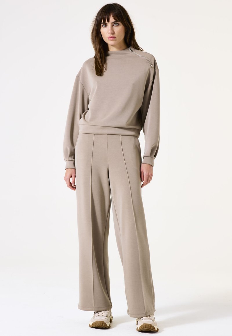 Beige sweatshirt en wijde broek, gemaakt van zachte stof; heeft een hoge halslijn en een zijritsdetail; gladde textuur overal.